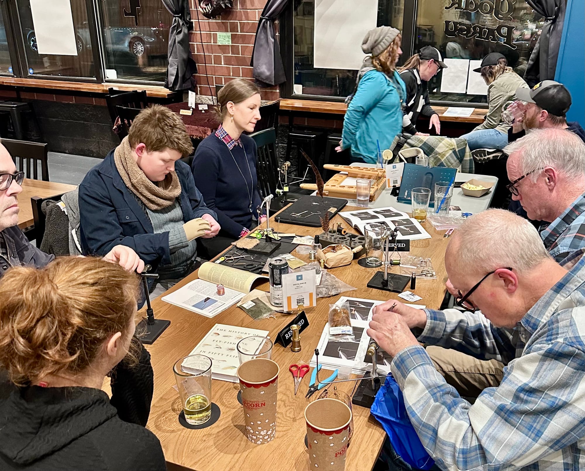 Group gathered around tables at fly tying night in a game bar, participants of various ages working at portable vises with materials and instruction sheets, beverages nearby
