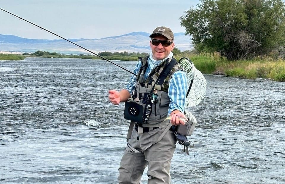 Fly fisherman in waders and vest standing in river with fly rod, mountains and trees in background under cloudy sky