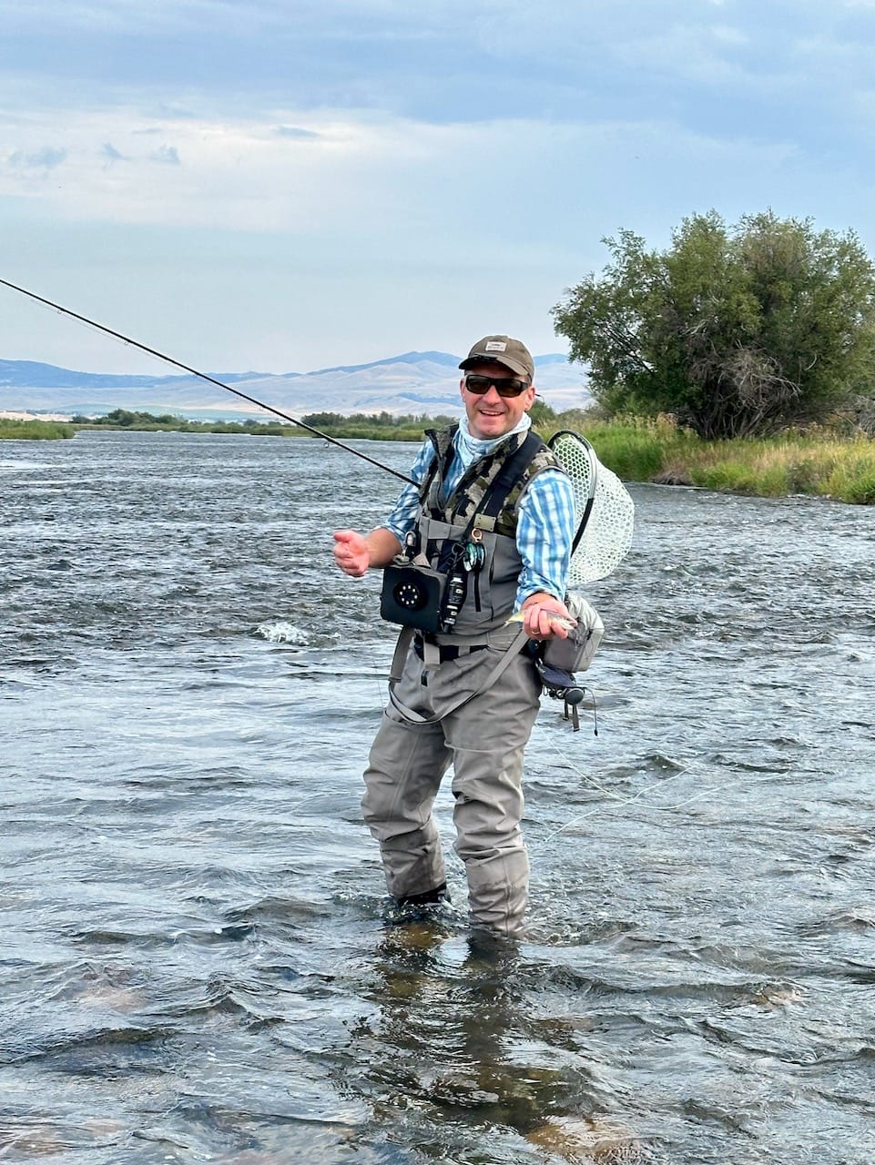 Fly fisherman in waders and vest standing in river with fly rod, mountains and trees in background under cloudy sky