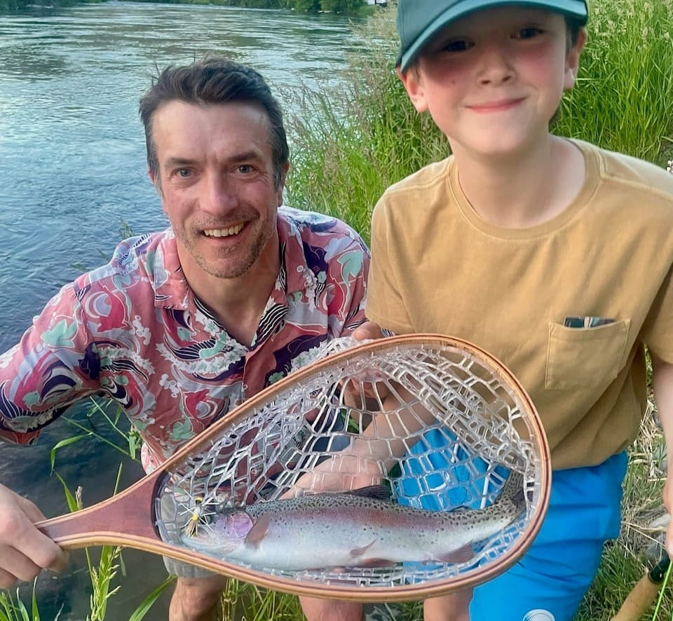 Adult and child holding rainbow trout in wooden net by the Deschutes river, child wearing Hardy cap, adult in colorful shirt