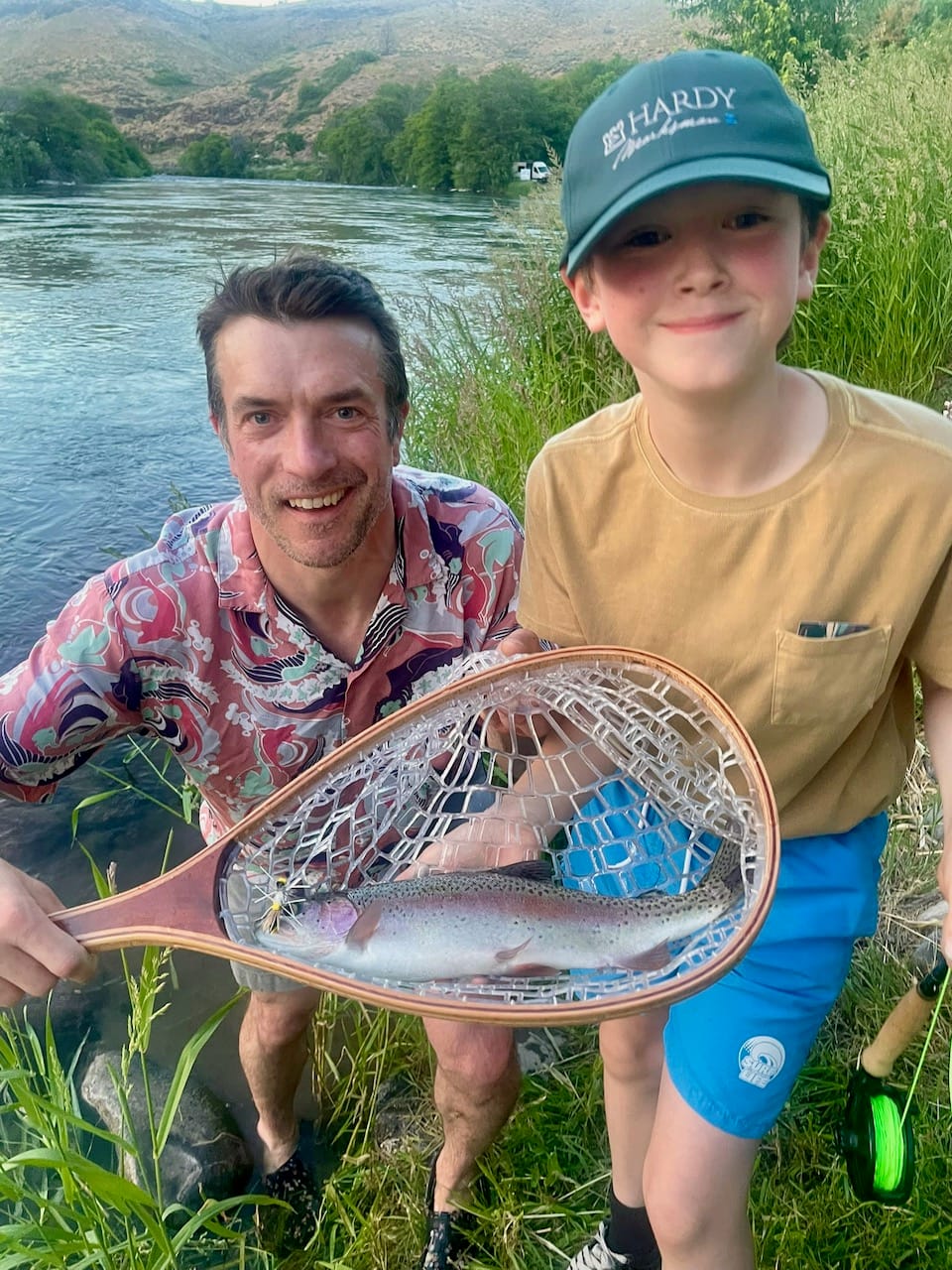 Adult and child holding rainbow trout in wooden net by the Deschutes river, child wearing Hardy cap, adult in colorful shirt
