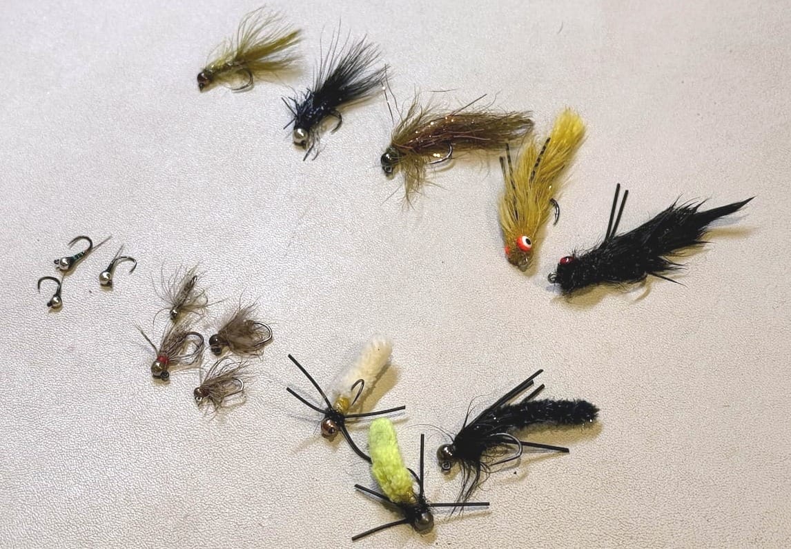 Assorted fly fishing flies laid out on white surface, ranging from small nymphs to mop flies to larger jig streamers