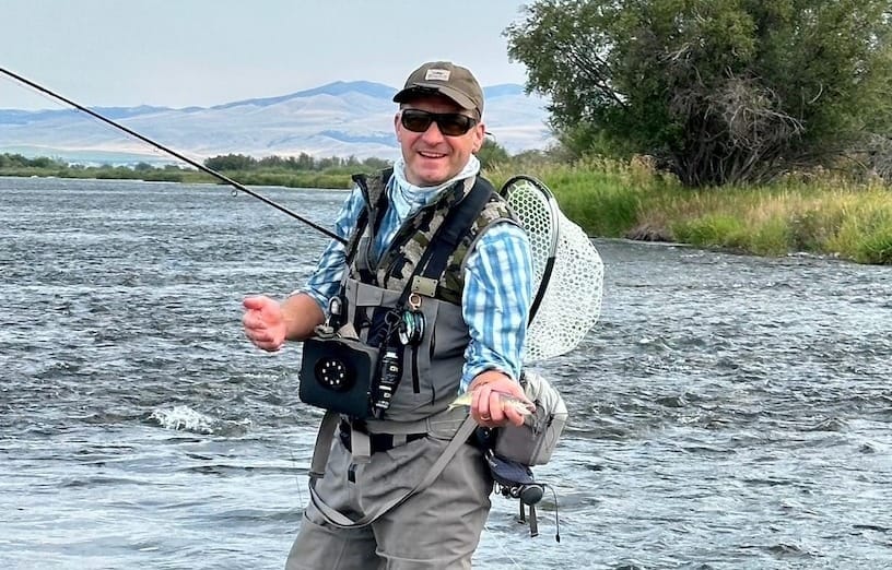 Smiling fly fisherman wading a Western river, holding a small trout with rod bent and net on back
