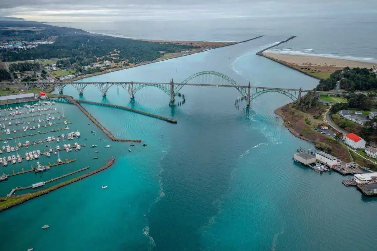 Aerial view of Yaquina Bay Bridge spanning an estuary in Newport, Oregon, turned teal from herring spawning, with a marina full of boats at left and the Pacific Ocean visible in the background.