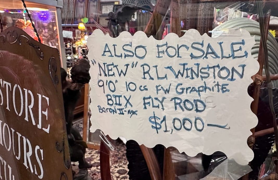 A handwritten sign in an antique shop window advertises a "new" R.L. Winston BIIX (Boron II-MX) 9'0" 10wt freshwater graphite fly rod for sale at $1,000.