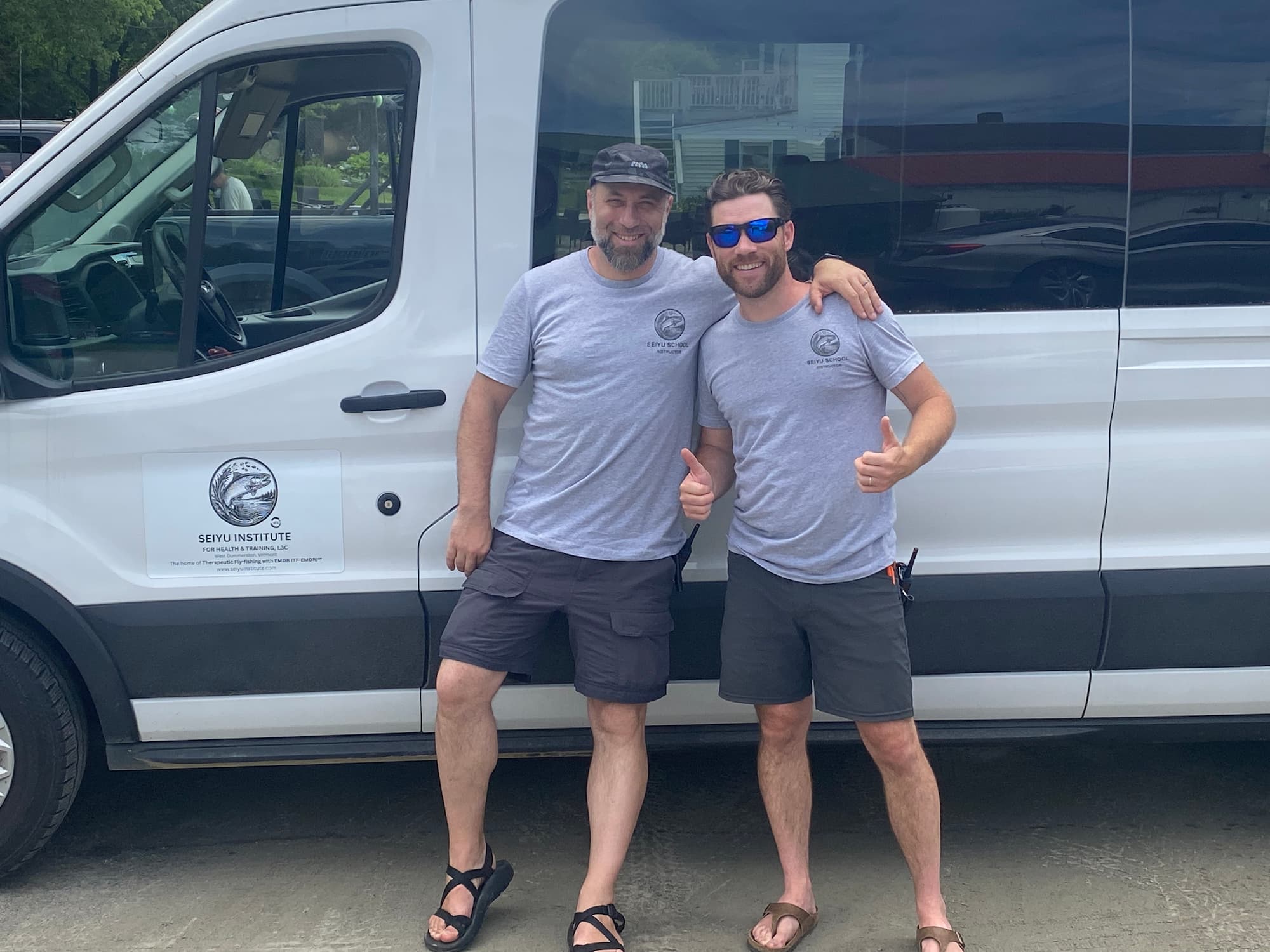 Two men in matching Seiyu Institute instructor shirts give thumbs up in front of a branded white Ford Transit van.