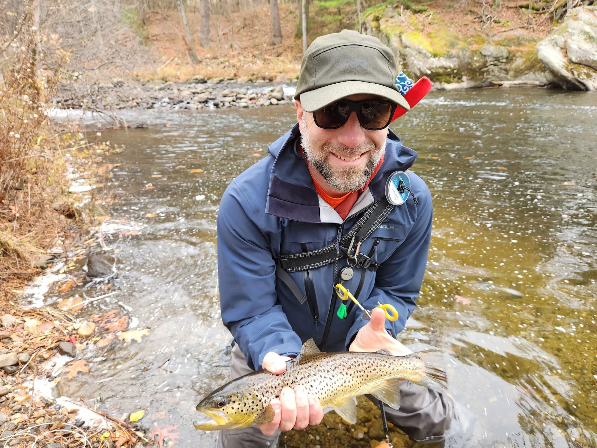Smiling angler holds a brown trout streamside in winter wading gear, fly reel visible on chest pack.