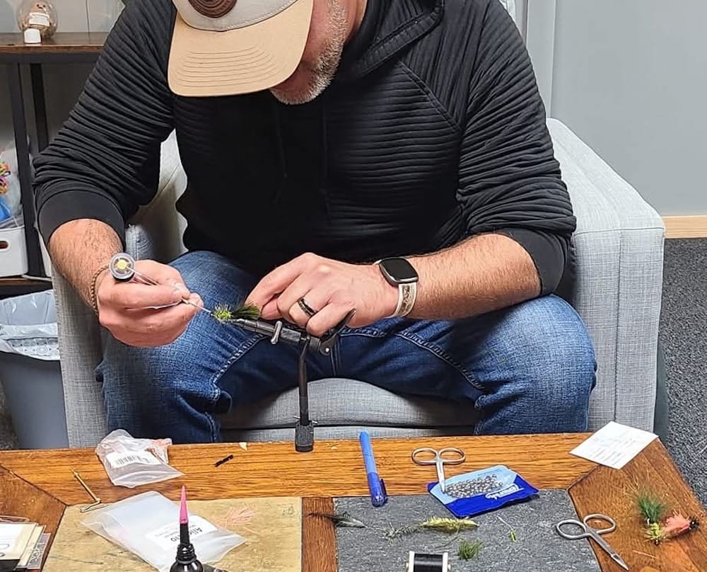 A man ties a fly during a group session, with tools and materials arranged on the table in front of him.