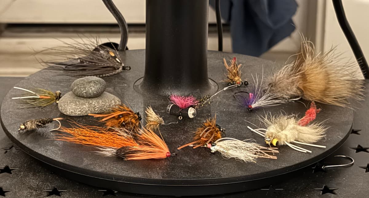 Assortment of fly fishing flies arranged on a monitor stand, including streamers, nymphs, and sculpins in varied colors.