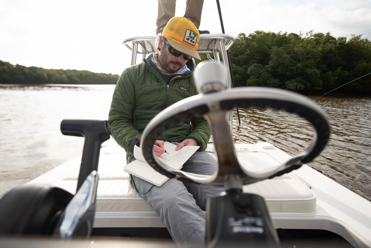 Angler in a yellow cap and green Patagonia jacket writing in a notebook while seated amidship in a flats boat on a calm estuary.