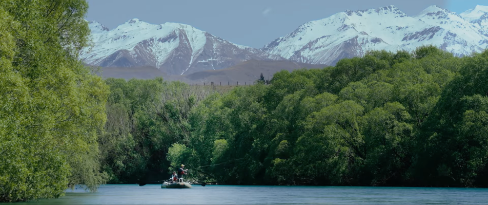 Two anglers fly fishing from a raft on a turquoise river, surrounded by lush green vegetation with snow-capped mountains rising dramatically in the background.