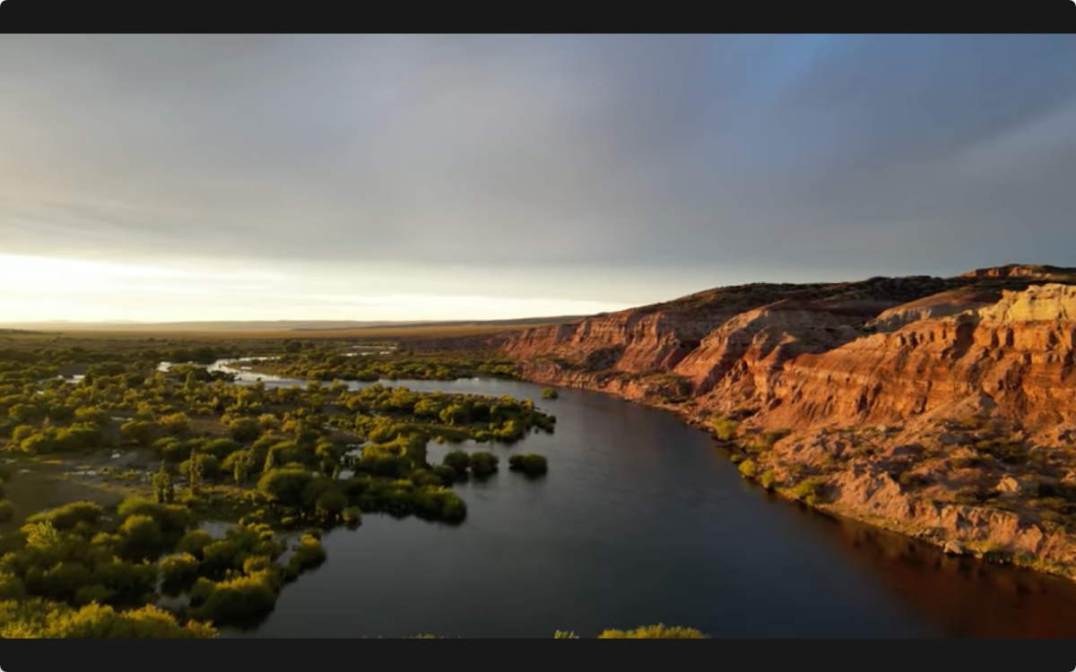 Aerial view of winding Limay Medio river through green valley with dramatic red rock cliffs at golden hour under stormy sky