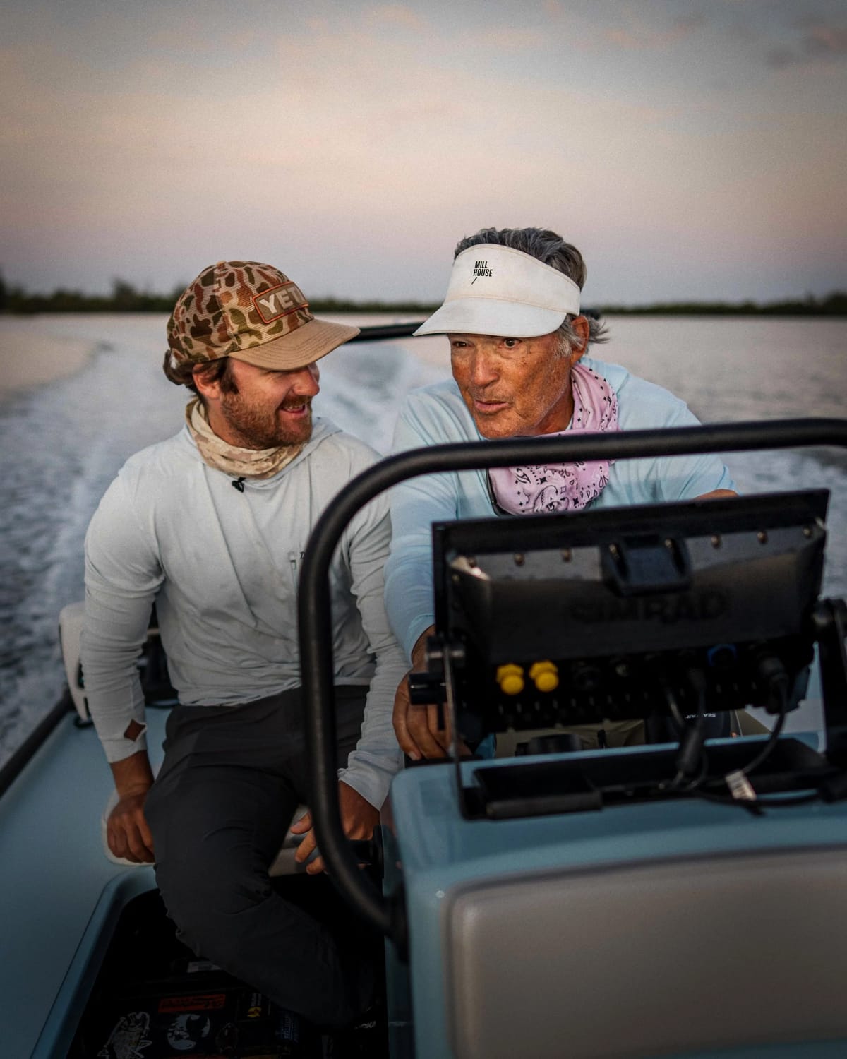 Two men, a younger and an older, at the console of a skiff boat tear-assing through the ocean are looking at each other. The older man drives the boat.