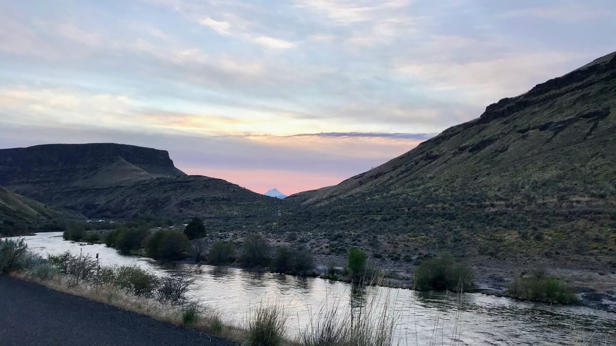 Scenic view of the Deschutes River winding through a desert canyon at sunset. The river reflects soft pink and purple twilight colors from the sky. 