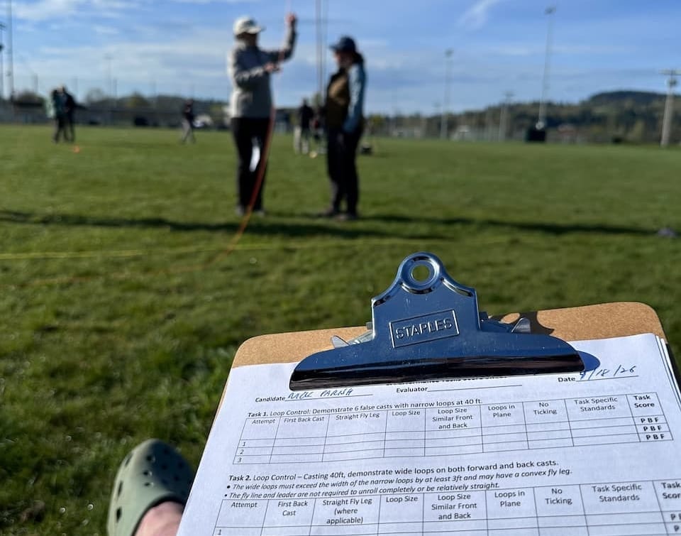 FFI Casting Instructor pre-test evaluation form on clipboard in foreground, an instructor and candidate learning fly casting on a grass field under partly cloudy skies in the background.