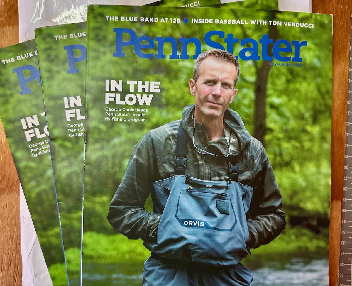 Three Penn Stater magazines fanned out; cover shows George Daniel in Orvis waders beside a stream, headlined "In the Flow: George Daniel leads Penn State's iconic fly-fishing program."