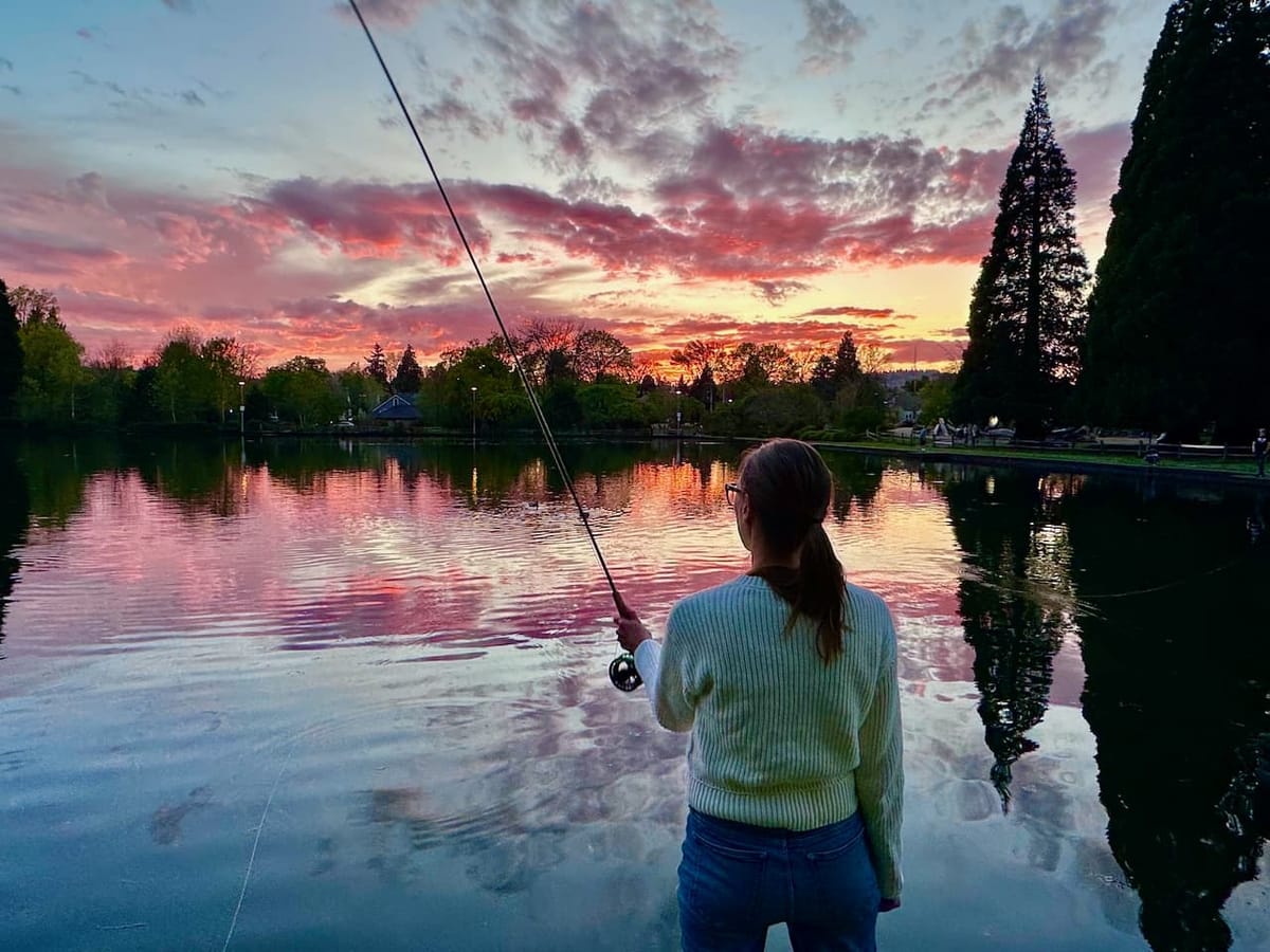 Woman practices roll cast on a calm pond at sunset, fly rod raised against a vivid pink and orange sky reflected in still water.