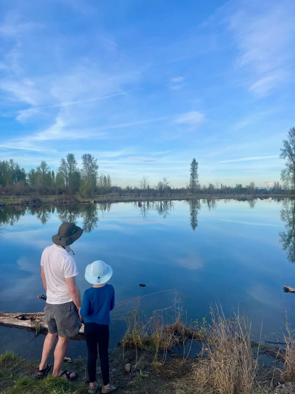 An adult and child stand by a pond where wispy clouds and far-off trees are reflected in the surface
