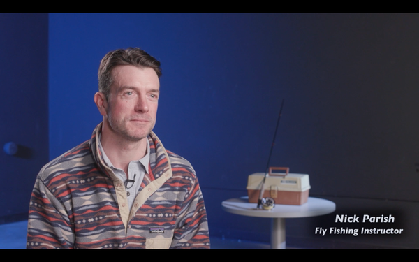 A curious-looking fellow in a patterned fleece sits foreground in a studio, with a tackle box and a fly rod in the background on a table. The super reads "Nick Parish Fly Fishing Instructor'
