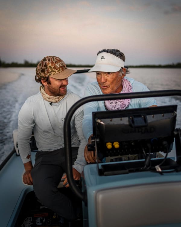 Two men, a younger and an older, at the console of a skiff boat tear-assing through the ocean are looking at each other. The older man drives the boat.