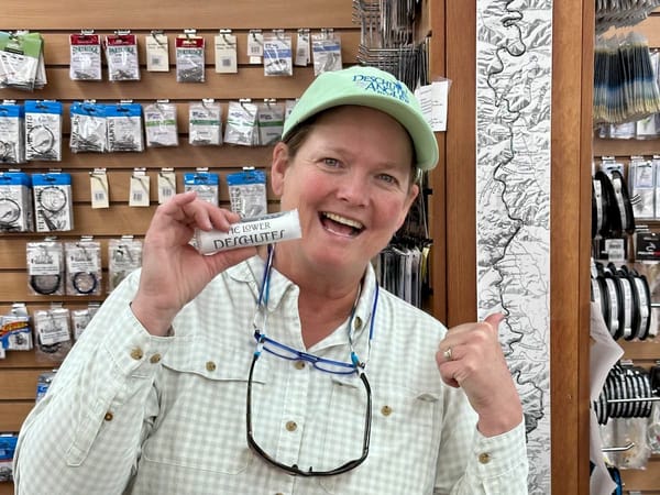 Amy Hazel, owner of the Deschutes Angler, holds up a rolled Lower Deschutes ribbon map inside her fly shop, grinning, pointing to the map behind her on the wall.