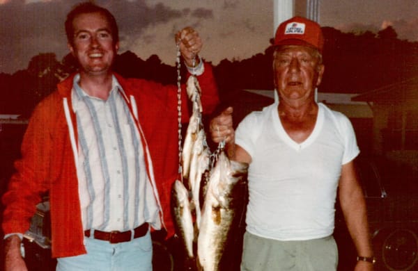 Two men posing at dusk with a stringer of fish including one gigantic largemouth bass, one in a red blazer and one in a white tee and red United Airlines cap, Florida, 1980s.