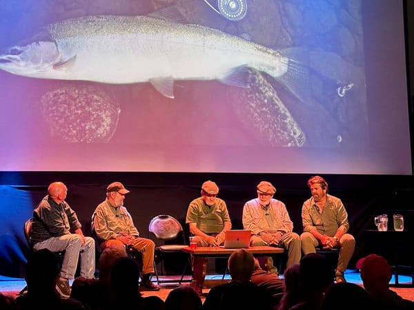 Five men sit on a theater stage looking at a photograph of a steelhead caught on a fly. It is a glorious fish.