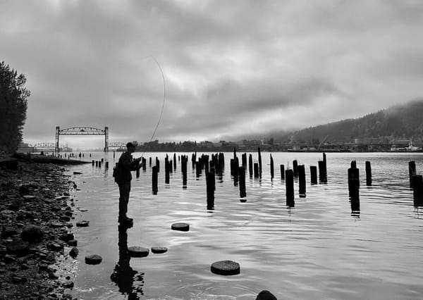 B&W photo of an angler casting a fly rod from a rocky riverbank amid wooden pilings, with a bridge in the cloudy distance.
